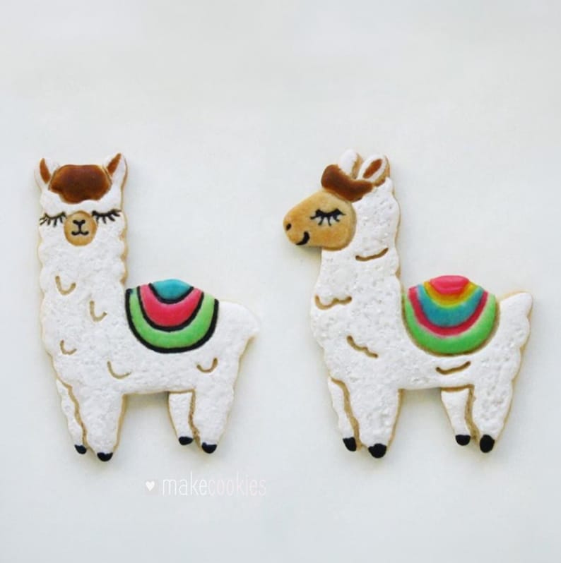 May include: Two llama-shaped cookies with white icing, brown ears, and colorful saddle blankets. The cookies have black feet and closed eyes. The text "makecookies" is at the bottom.