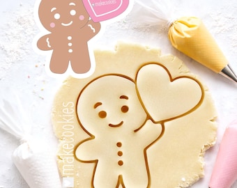 Könnte beinhalten: Ausstecher in Form eines Lebkuchenmanns, der ein herzförmiges Schild mit dem Text "makecookies" hält. Der Ausstecher ist in einen Teig gedrückt, mit einem passenden Aufkleber. Spritzbeutel sind im Hintergrund.
