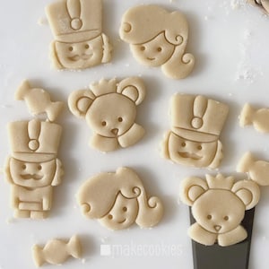 May include: A collection of unbaked cookie dough cutouts in various shapes, including a soldier, a princess, a bear, and candies. The cookies are light beige in color and arranged on a white surface. The image includes the text "makecookies".
