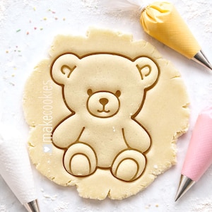 May include: A light-colored, bear-shaped cookie cutter pressed into dough. The cutter has a detailed outline of a teddy bear. The image includes the text "makecookies" and several pastry bags with metal tips.