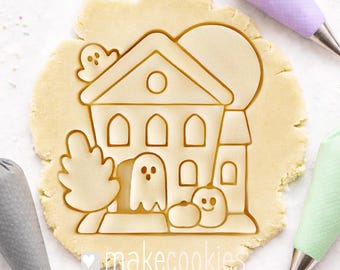 May include: A Halloween-themed cookie cutter pressed into dough. The design features a haunted house, ghosts, a tree, and a smiling pumpkin. The text "makecookies" is visible at the bottom. Icing bags in gray, purple, and green are in the background.