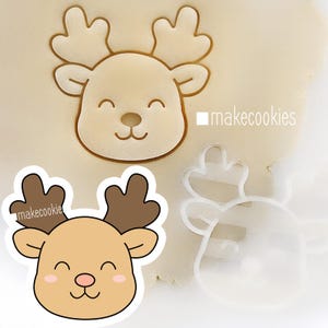 May include: A reindeer cookie cutter with a smiling face and antlers is pressed into dough. A finished cookie and a line drawing of the reindeer are also shown. The text "makecookies" is visible.