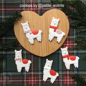 May include: Five llama-shaped cookies with white frosting, red accents, and patterned scarves. The cookies are arranged on a wooden heart-shaped board and a plaid background. The text "cookies by @petite_events" is visible.