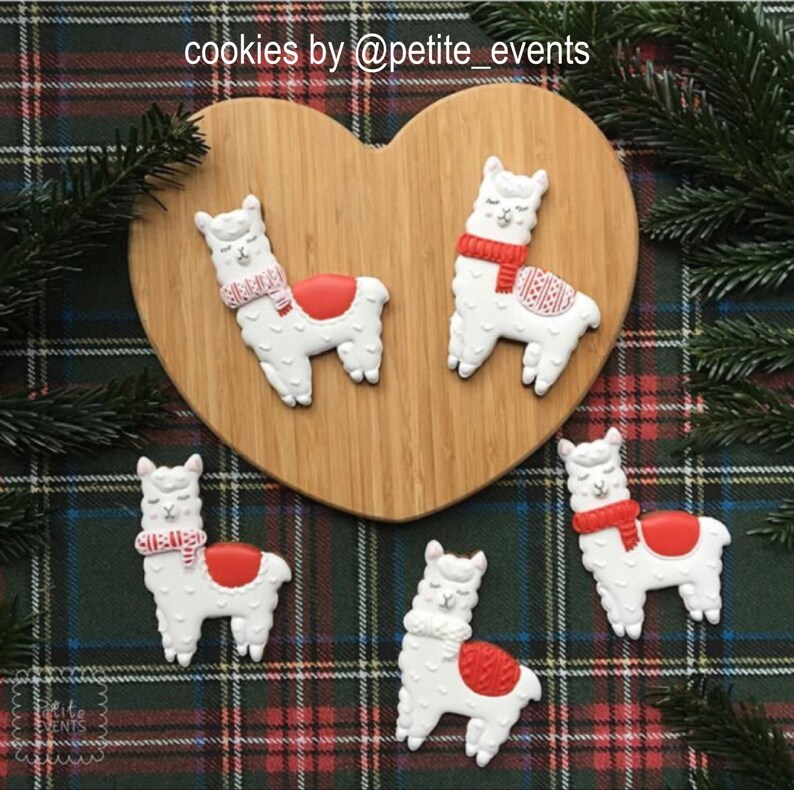 May include: Five llama-shaped cookies with white frosting, red accents, and patterned scarves. The cookies are arranged on a wooden heart-shaped board and a plaid background. The text "cookies by @petite_events" is visible.