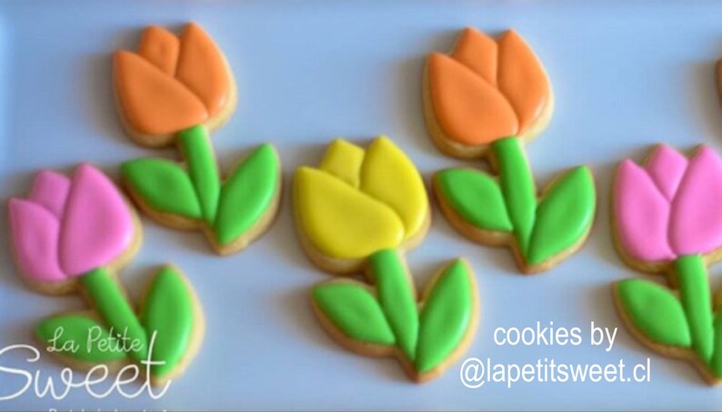 Op de afbeelding: Vijf gedecoreerde suikerkoekjes in de vorm van tulpen. De koekjes zijn versierd met roze, oranje en gele glazuur. De bladeren zijn groen. De tekst "La Petite Sweet" staat in de linkerbenedenhoek van de afbeelding. De tekst "cookies by @lapetitsweet.cl" staat in de rechterbenedenhoek van de afbeelding.