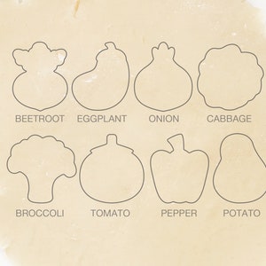May include: A set of eight cookie cutters shaped like different vegetables, including a beet, eggplant, onion, cabbage, broccoli, tomato, pepper, and potato. The cutters are arranged on a white surface.