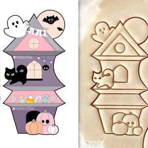 May include: A Halloween-themed cookie cutter in the shape of a haunted house. The house is purple, pink, and gray, with a black cat, ghost, and pumpkins. The word "BOO" is on a banner. The cookie cutter is for making Halloween treats.