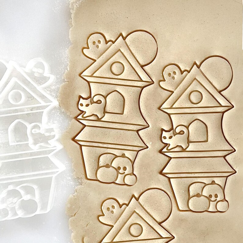 May include: Cookie cutter with a Halloween theme. The design includes a haunted house with a cat, ghost, and pumpkin. The cutter is made of clear plastic and is next to cookies cut with the same design. Perfect for making Halloween cookies.