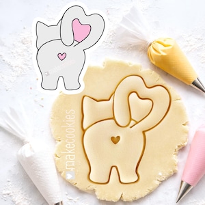 May include: A cat-shaped cookie cutter with a heart-shaped tail and a heart on its rear. The cutter is next to a cutout in dough, with piping bags in the background. The text "makecookies" is visible.