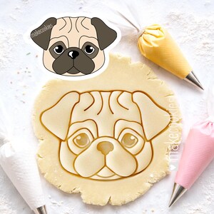 May include: A pug-shaped cookie cutter and a pug-shaped cookie cut-out in dough. The cookie cutter is beige, brown, and black. The image also shows piping bags with yellow and pink frosting.