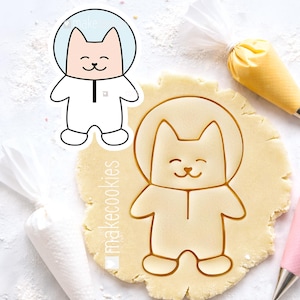 Puede incluir: Un cortador de galletas con forma de gato y una pegatina a juego. El gato lleva un casco y un traje de astronauta. El cortador de galletas está presionado en una masa. La pegatina y el cortador de galletas tienen el texto "makecookies".