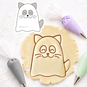 May include: A cat-shaped cookie cutter with a ghost-like design, stamped into a circle of dough. The cutter has a smiling face and the text "makecookies". Three pastry bags with metal tips are in the background.
