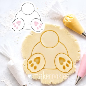 May include: A bunny butt cookie cutter with a matching cutout in dough. The cutter is white with pink paw pads. The dough is light brown. The image also includes piping bags with frosting.