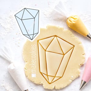 May include: A geometric crystal-shaped cookie cutter pressed into dough, with a sticker of the same design. Piping bags are placed around the dough. The text "makecookies" is visible on the dough.