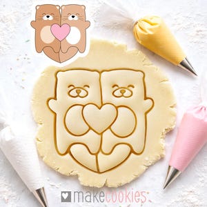 Puede incluir: Un cortador de galletas con la forma de dos nutrias abrazando un corazón, junto a un corte en la masa. Las nutrias son marrones con vientres beige y un corazón rosa. La masa presenta el mismo diseño de nutria y corazón. Hay mangas pasteleras en el fondo.