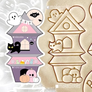 May include: A Halloween-themed cookie cutter in the shape of a haunted house. The house is purple, pink, and gray, with a black cat, ghost, and bat. The text "makecookies" is visible. The cookie cutter is next to a cookie with the same design.