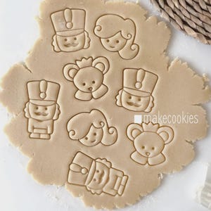 May include: Cookie dough with impressions of a nutcracker, a princess, and a bear with a crown. The cookie cutters are pressed into the dough, creating detailed shapes. The text "makecookies" is visible on the dough.