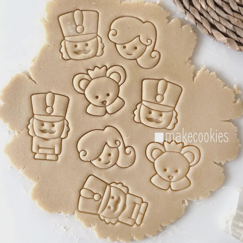 May include: Cookie dough with impressions of a nutcracker, a princess, and a bear with a crown. The cookie cutters are pressed into the dough, creating detailed shapes. The text "makecookies" is visible on the dough.