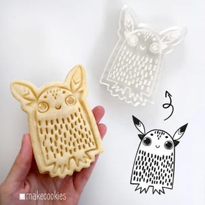 May include: A cookie cutter and a baked cookie in the shape of a whimsical creature with large eyes and pointed ears. The cookie cutter is clear plastic, and the cookie is golden brown. A black and white illustration of the creature is also shown.