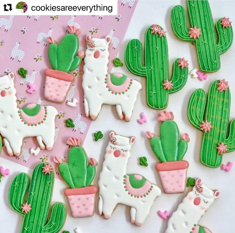 May include: An assortment of decorated sugar cookies featuring a llama and cactus theme. The cookies are white, green, pink, and brown. The llamas have pink and green saddle blankets and flower accents. The cacti are in pink pots. The background is pink with llama illustrations.