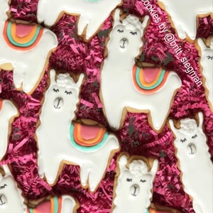 May include: Close-up of several llama-shaped cookies with white icing and colorful saddle blankets. The cookies are decorated with black eyes and eyelashes. The cookies are arranged on a bed of pink shredded paper.