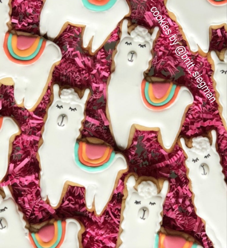 May include: Close-up of several llama-shaped cookies with white icing and colorful saddle blankets. The cookies are decorated with black eyes and eyelashes. The cookies are arranged on a bed of pink shredded paper.