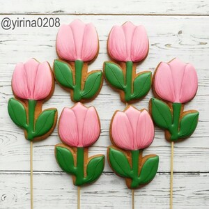 Op de afbeelding: Zes roze en groene suikerkoekjes versierd om eruit te zien als tulpen. De koekjes zitten op stokjes en zijn gerangschikt op een wit houten oppervlak.