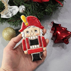 May include: A hand holding a ceramic Christmas nutcracker ornament. The ornament is painted with a red hat and coat, black pants, and a cream-colored face. A red Christmas tree ornament and a gold glitter ball are also visible.