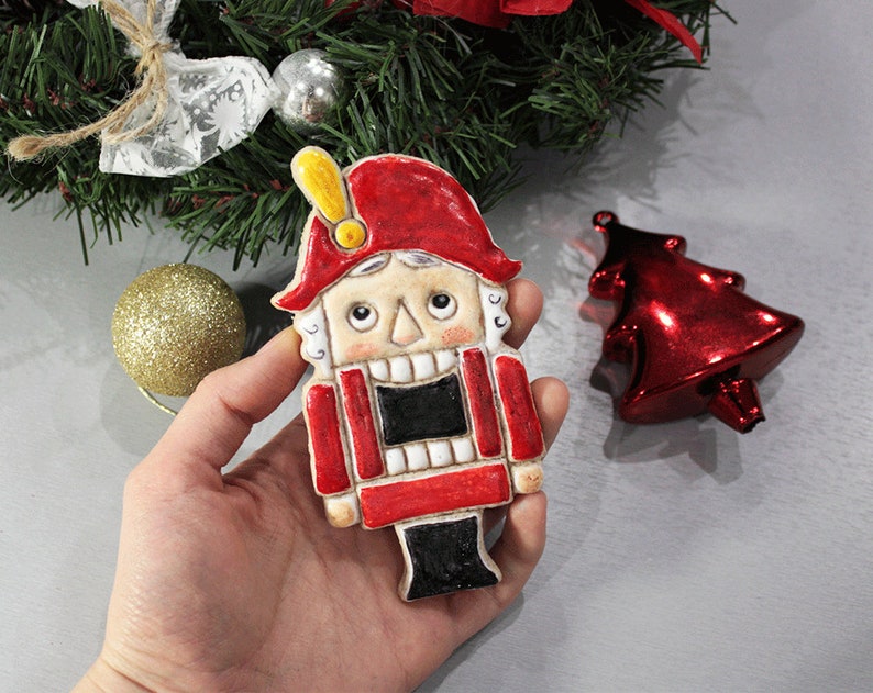 May include: A hand holding a ceramic Christmas nutcracker ornament. The ornament is painted with a red hat and coat, black pants, and a cream-colored face. A red Christmas tree ornament and a gold glitter ball are also visible.