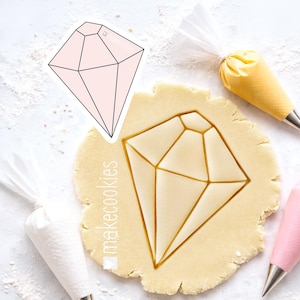 May include: A diamond-shaped cookie cutter and a cut-out in dough. The cutter is pink with black outlines, and the dough is light brown. Icing bags in white, yellow, and pink are also present. The text "makecookies" is visible.