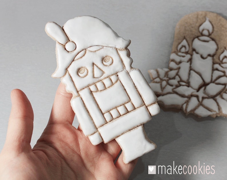 May include: A white, detailed cookie in the shape of a nutcracker soldier is held in a hand. The cookie has a hat, a square body, and a detailed face. Another cookie, shaped like candles and poinsettias, is in the background. The logo "makecookies" is in the bottom right corner.
