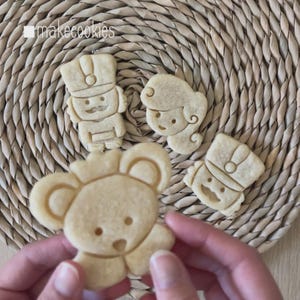 May include: Close-up of several freshly baked cookies. The cookies are shaped like a koala, a soldier, and a girl. The cookies are light brown in color and have detailed features. The cookies are on a woven mat. The text "makecookies" is in the top left corner.