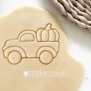 May include: A cookie cutter in the shape of a truck carrying a pumpkin, pressed into dough. The cutter is light brown, and the dough is a pale yellow. The text "makecookies" is printed in white below the truck.