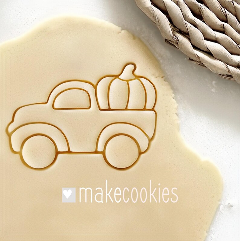 May include: A cookie cutter in the shape of a truck carrying a pumpkin, pressed into dough. The cutter is light brown, and the dough is a pale yellow. The text "makecookies" is printed in white below the truck.