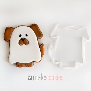 Op de afbeelding: Een witte spookkoekje met bruine oren, een bruine neus en bruine voeten. Het koekje ligt naast een doorzichtige plastic koekjesuitsteker in de vorm van een spookhond.