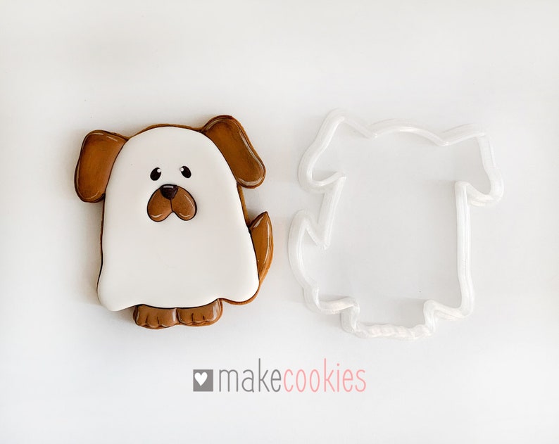 Op de afbeelding: Een witte spookkoekje met bruine oren, een bruine neus en bruine voeten. Het koekje ligt naast een doorzichtige plastic koekjesuitsteker in de vorm van een spookhond.