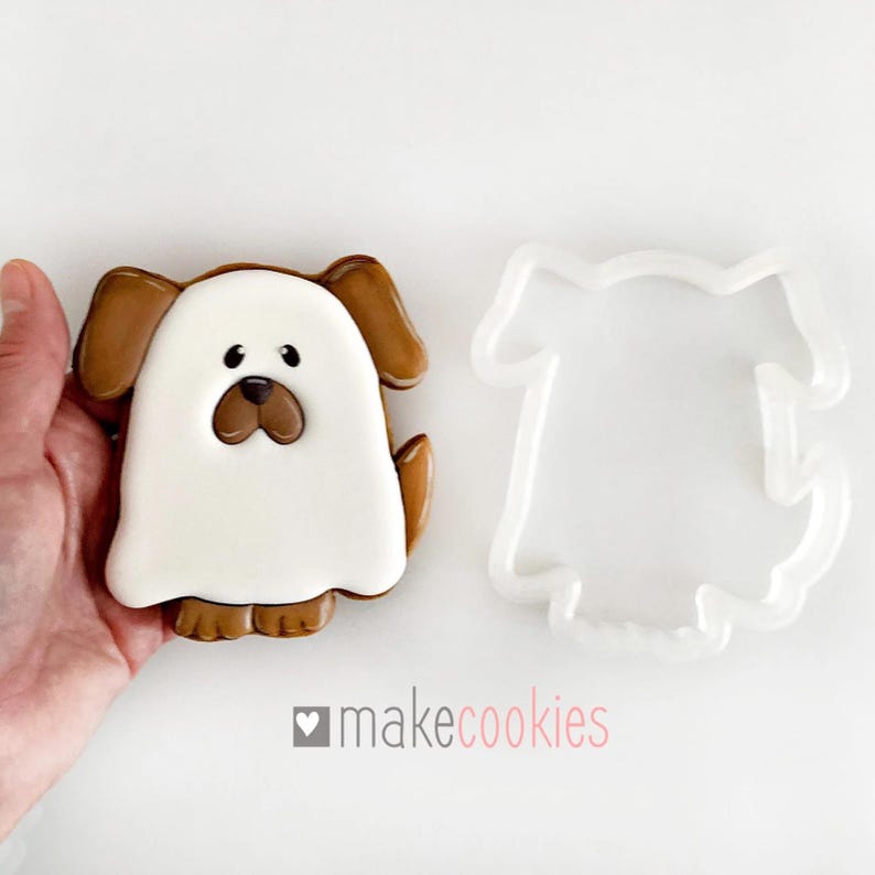 Op de afbeelding: Een witte en bruine spookhond koekjesvormer met een bruine omtrek en een wit spookachtig lichaam. De koekjesvormer wordt getoond naast een gedecoreerde koek in de vorm van de vormer.