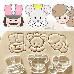 May include: Set of six cookie cutters, including a nutcracker, a bear, a princess, and three clear plastic cutters. The cutters are on a brown surface. The text "makecookies" is visible in the image.