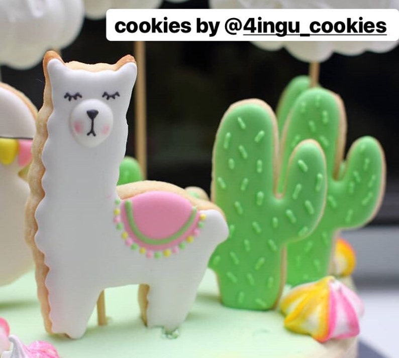 May include: Cookies decorated with white icing in the shape of a llama and green cactus. The llama cookie has a pink saddle and the cactus cookies have green icing with small green sprinkles. The text "cookies by @4ingu_cookies" is at the top.