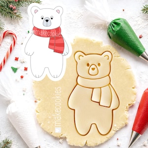 May include: A white polar bear cookie cutter with a red scarf, alongside a bear-shaped cookie cut into dough. The cookie cutter has the text "makecookies". Festive baking supplies are in the background.