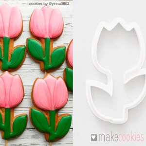 Op de afbeelding: Zes roze en groene suikerkoekjes in de vorm van tulpen, versierd met roze en groene glazuur. Naast de koekjes is een witte plastic koekjesuitsteker in de vorm van een tulp te zien. De tekst "makecookies" staat rechtsonder in de afbeelding.