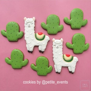 May include: Cookies decorated as white llamas with colorful saddle blankets and green cacti on a pink background. The cookies are arranged in a visually appealing pattern. The text "cookies by @petite_events" is at the bottom.