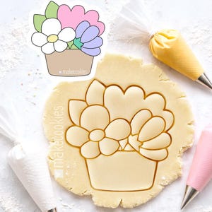 May include: A floral cookie cutter with a bouquet design. The cutter is in the shape of a basket filled with flowers in white, pink, and purple. The text "makecookies" is printed on the cutter. The cutter is on a surface of cookie dough, with piping bags in the background.