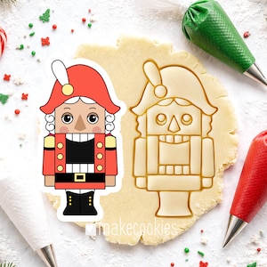 May include: A red, black, and gold nutcracker cookie cutter and a cookie cut-out on a light brown surface. The nutcracker has a red hat, a black uniform, and a white face. A green and a red piping bag are in the background.
