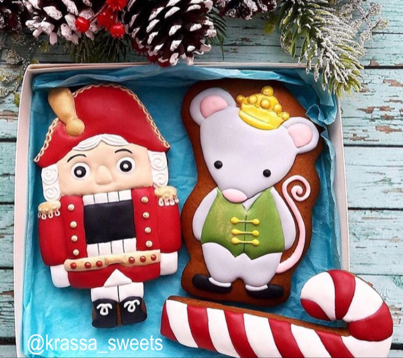 May include: A box of decorated gingerbread cookies featuring a Nutcracker soldier in a red coat and a mouse wearing a crown and vest. A red and white candy cane is also included. The cookies are on a blue background with the text "@krassa_sweets".