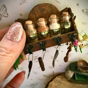 Puede incluir: Un estante de especias de madera en miniatura con cuatro frascos de vidrio etiquetados como "Sal", "Pimentón", "Pimienta" y "Sésamo". El estante tiene cinco ganchos colgando, cada uno con un pequeño objeto de metal.