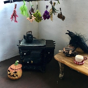 May include: A miniature dollhouse scene featuring a black cast iron stove, a small wooden table with books and a teacup, a jack-o'-lantern, and a black raven. A branch hangs from the ceiling with dried flowers and herbs.