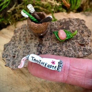 以下が含まれることがあります： 赤い花と「Toothpaste」の文字が書かれた、ミニチュアの白い歯磨き粉のチューブ。チューブは指の上に置かれています。