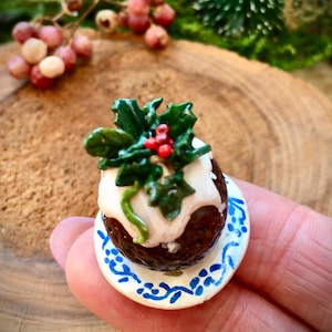 Könnte beinhalten: Ein Miniatur-Weihnachtspudding mit weißem Zuckerguss und einem grünen Stechpalmenzweig obenauf, der auf einem kleinen Teller mit blau-weißem Muster steht.
