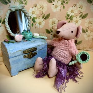 May include: A handmade pink mouse doll wearing a pink sweater and purple scarf, seated next to a blue wooden box and a small mirror. The doll holds a small green hand mirror. A small vanity set is on the box.